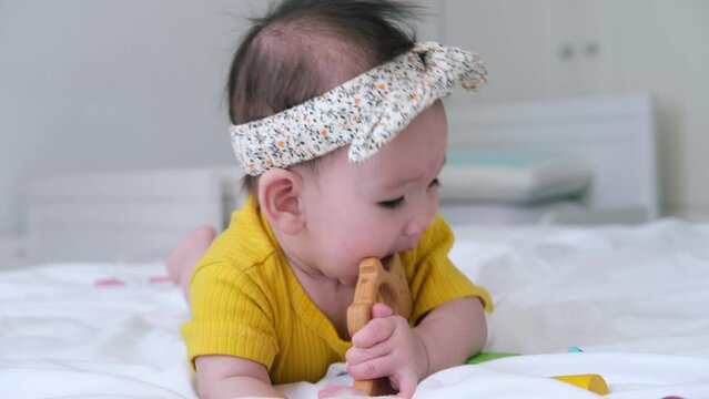 healthy asian baby girl in yellow bodysuit plays with wooden toy teether for teeth on white bedding