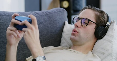 Phone, gaming and headphones with a man on a sofa in the living room of his home to play a mobile game. Internet, technology and video games with a male gamer nerd playing online to relax in a house