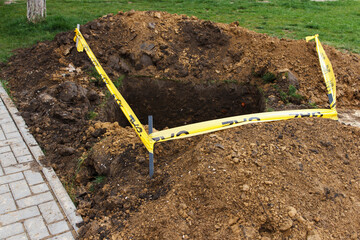 A square pit dug into the soil, next to public sidewalk and fenced with a ribbon, as a sign of danger. Communal service works, eliminate problems underground
