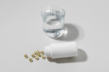 Yellow pill capsules falling out of a white blank jar near a transparent glass of water on a gray isolated background. Concept of pharmacy, drugs for health and energy. Image for your design