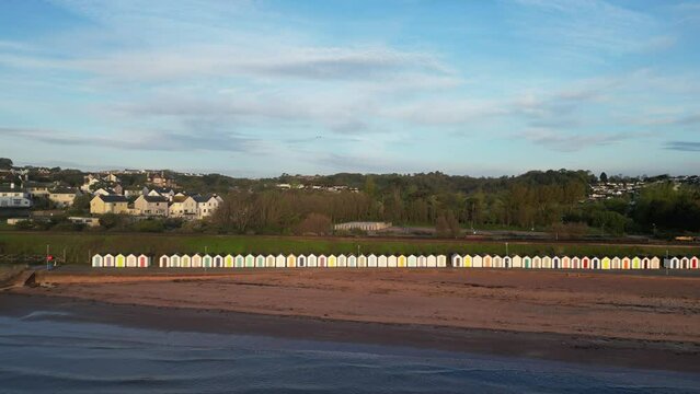 Goodrington Sands, Torbay, South Devon, England: DRONE AERIAL VIEWS: The drone flies in over the Torbay sea and Goodrington Sands beach towards a long row of multi-coloured beach huts at dawn.
