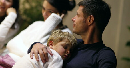 Young father holding sleeping child at night while in conversation with friends, candid casual man with son