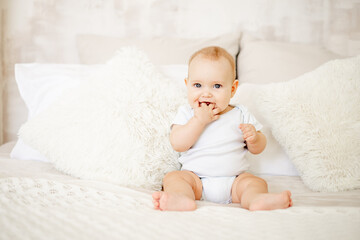 a baby boy or girl of six months is sitting at home on a bed in a bright bedroom and smiling or laughing, a happy newborn in a white bodysuit