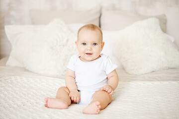 a baby boy or girl of six months is sitting at home on a bed in a bright bedroom and smiling or...