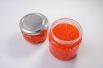 Open glass jar of red trout fish caviar on a white background.