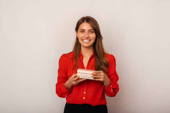 Ecstatic woman wearing red shirt is holding a small gift box on grey background.