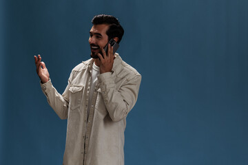 Portrait of a man brunette animation and joy talking on the phone hand up from surprise victory and happiness smile with teeth, on a blue background in a white T-shirt and jeans, copy space