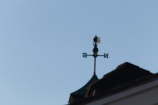 This weather vane was beautiful on the roof contrasted against the sky. The shadowy image with the clear blue sky in the background. The North, South, East, and West showing direction of the wind.