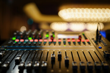 Faders pushed up on audio mixer console with LED buttons in bokeh in well lit ballroom.. Lights are blurred for effect.