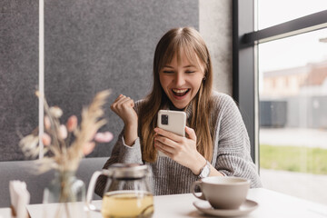Joyful blonde woman playing video game on mobile phone, reading good news, celebrating winning...