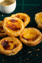 Group of traditional Portuguese pastel de nata on cooling wire rack and cup of espresso on green tiled surface.