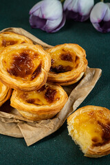 Pile of traditional Portuguese pastel de nata on paper bag on green surface with purple tulips on background.