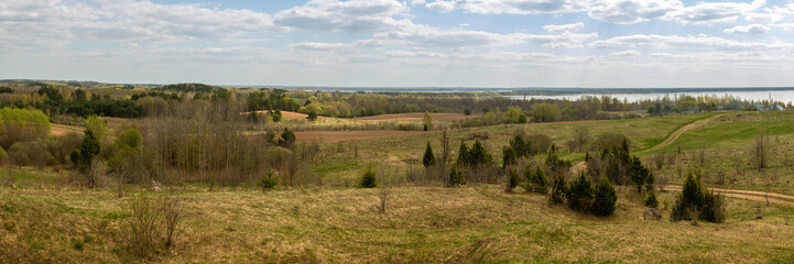 wide panoramic view of a hilly field with wild grass and trees, farmland, forest belts and a lake on a spring day under a blue cloudy sky