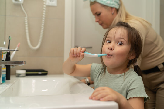 A little girl brushes her teeth and makes faces in the bathroom, her mother is in the background.