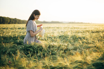 Naklejka na ściany i meble Beautiful woman in floral dress walking in barley field in sunset light. Atmospheric tranquil moment, rustic slow life. Stylish female holding straw hat and enjoying evening summer countryside