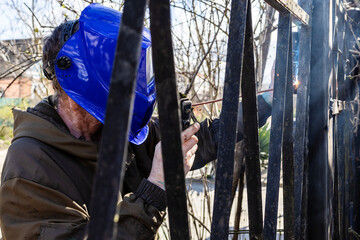 welder welds door hinge on gate of fence in village outdoors