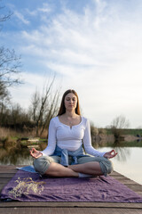 Fototapeta na wymiar Woman relaxingly practicing meditation in nature by the lake. Nature background. Spiritual and emotional concept.