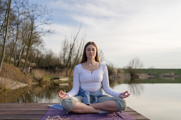 Fototapeta na wymiar Woman relaxingly practicing meditation in nature by the lake. Nature background. Spiritual and emotional concept.