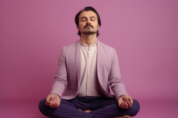Young man meditating in lotus position while sitting on floor against purple background