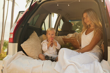 Young pretty woman in white dress and her little son sitting in car trank in nest of cushions, boy eating apple, family enjoying summer day, happy holiday concept