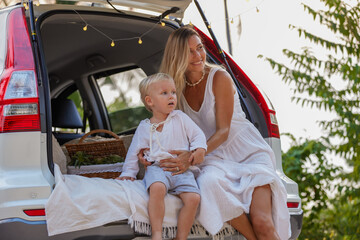 Happy young mother in white clothes with baby son sitting in open car trunk having picnic outdoors, family enjoying summer day in park, family time concept, copy space