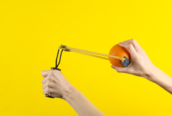 Hands holding slingshot with orange on a yellow background