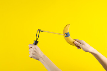 Hands holding slingshot with banana on a yellow background - obrazy, fototapety, plakaty