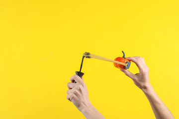 Hands holding slingshot with toy pumpkin on a yellow background. Halloween theme, trick or treat