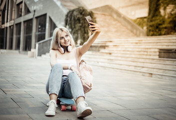 Young smiling cool girl sitting on skateboard and makes selfie on phone in the city