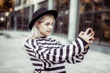 Young charismatic smiling woman in hat makes selfie on phone outdoors