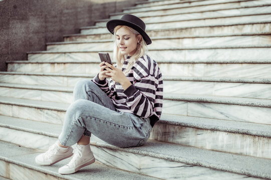 Young charming hipster girl in hat using phone while sitting on the stairs outdoors