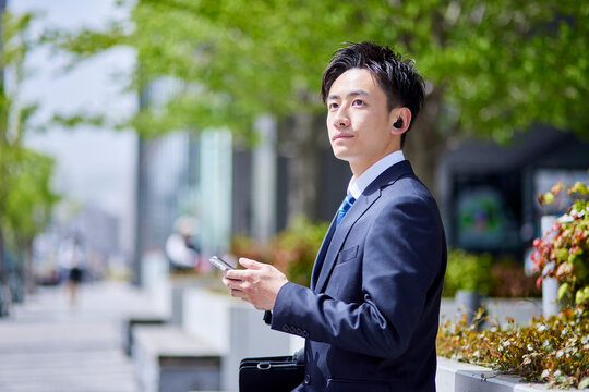 20代日本人ビジネスマンがワイヤレスイヤホンとスマートフォンを使用して仕事をしている春のイメージ