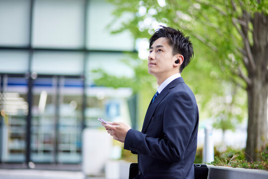 20代日本人ビジネスマンがワイヤレスイヤホンとスマートフォンを使用して仕事をしている春のイメージ
