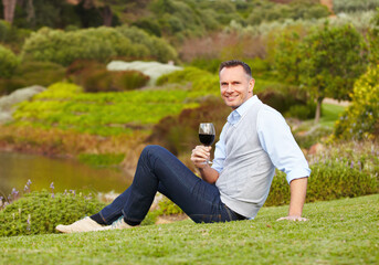 Mature man with red wine outdoor, relax with smile in nature with alcohol drink, leisure and travel to countryside. Happy male person with alcoholic beverage in glass, satisfied and relaxing in park