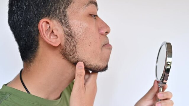 Asian man looking and touching his beard grows on a part of his lower face by mini mirror. Adult men typically have thousands of beard hairs.