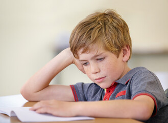 School, education and a boy reading a book at his desk in a classroom for studying or child development. Kids, learning and bored with a young male student child in class to study for an exam or test