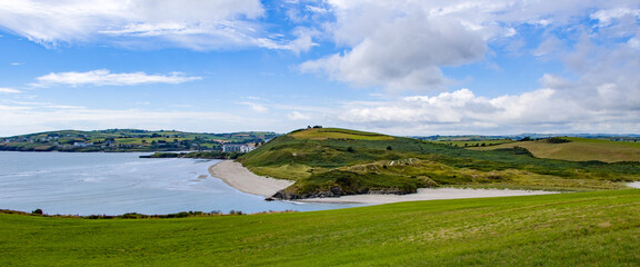 The green hills of Inchydoney Island and the famous Irish beach on a sunny summer day. Clear sky with white clouds over the Irish coast. Irish summer landscape. Green grass field near water