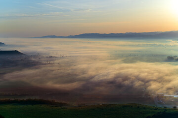 Aerial view of the valley in early morning mist, beautiful in the highlands. Low clouds and fog cover the sleeping meadow. Alpine mountain valley mists landscape at dawn. Serene moment in rural area