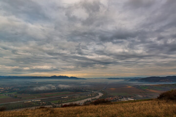 Naklejka na ściany i meble Serene moment in rural area in beautiful early morning mist. Panoramic view of a sleeping meadow valley covered with low clouds and fog, a highway crossing a river, rolling hills and villages