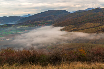 Serene moment in rural area in beautiful autumn early morning mist. Panoramic view of a sleeping meadow valley covered with low clouds and fog, village hiding between rolling hills covered with trees