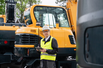 Engineer in a helmet with a digital tablet stands next to construction excavators