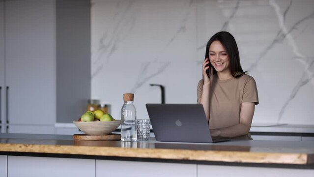 Young  woman working on laptop answering phone call in the kitchen