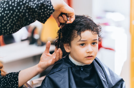Little curly hispanic boy sitting at barber shop looks away. Cropped shoot of woman hairdresser making haircut fir male toddler. Children beauty, childhood.