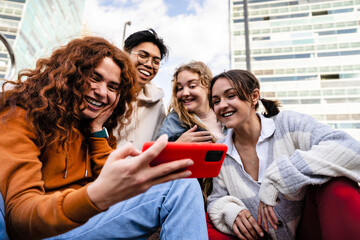 Group of young gen z friends sitting together in the city using cell phone app to share funny content - obrazy, fototapety, plakaty