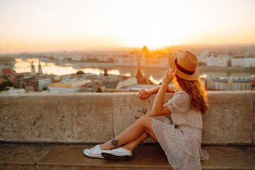 Beautiful woman in hat enjoys stunning view of the city at dawn. Back view. Tourist is looking at...