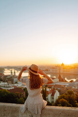 Beautiful woman in hat enjoys stunning view of the city at dawn. Back view. Tourist is looking at...