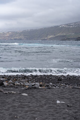 Black sand beach with waves. Breakwater on one side. Cliffs with houses in the background. Stones in the sand. Cloudy day. Puerto de la Cruz, Tenerife, Canary Islands, Spain.