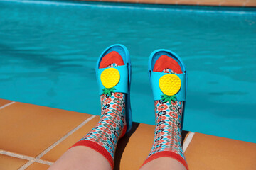 Feet with colorful socks and blue pool sandals at the edge of a swimming pool. Summer concept....