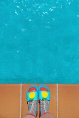 A young woman's feet in colorful socks and blue flip-flops at the edge of a swimming pool. Summer...