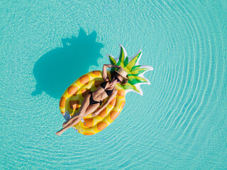 Sexy young female in swimsuit bikini, in a straw hat and sunglasses floating on blue swimming pool waves on giant inflatable Yellow Pineapple tube. Chill out summer vacation in luxury resorts concept
